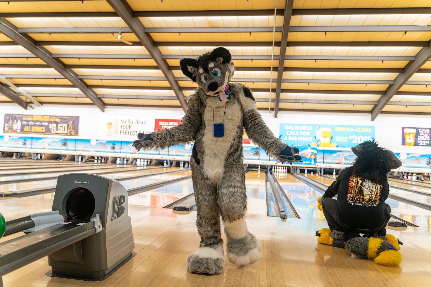 A grey fursuit character stands with arms outstretched on the bowling lanes.