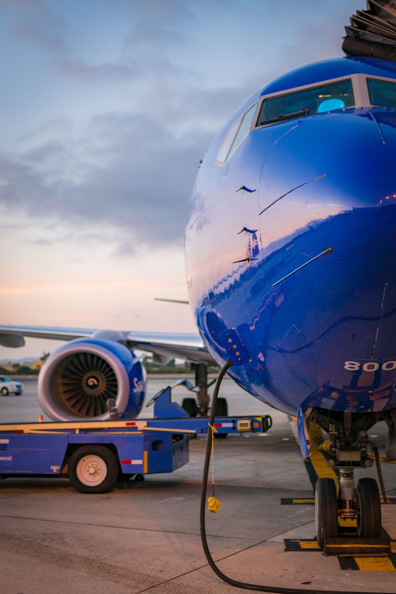Blue commercial aircraft nose and fuselage with engine visible during sunset at an airport gate.