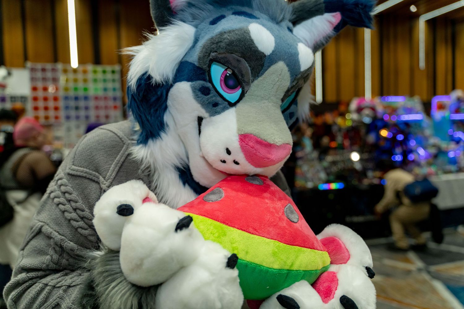 A furry mascot character with blue and white fur holds a colorful watermelon slice at what appears to be a convention.