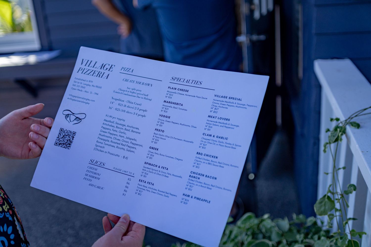 Hand holding menu from Village Pizzeria against blue building exterior.