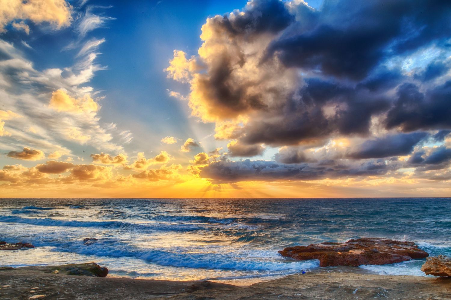 Dramatic coastal sunset panorama with stormy clouds and crashing waves against rocky shore.