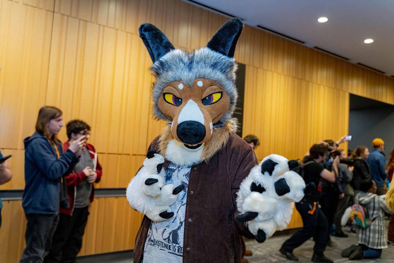 Furry mascot characters posing and interacting with attendees at a convention in a wood-paneled venue.