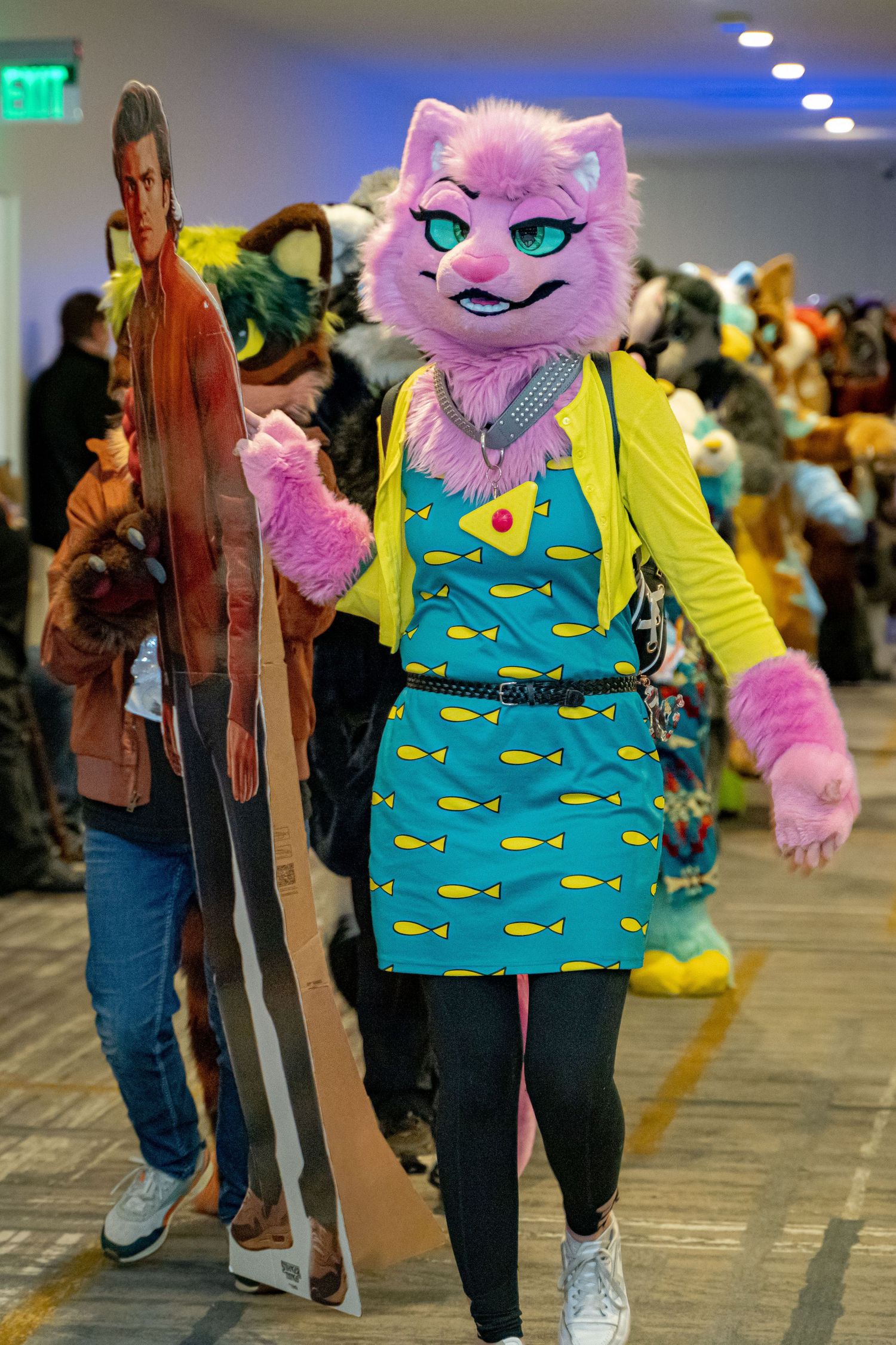 A person in a pink furry costume with a blue and yellow patterned vest poses playfully at a convention.