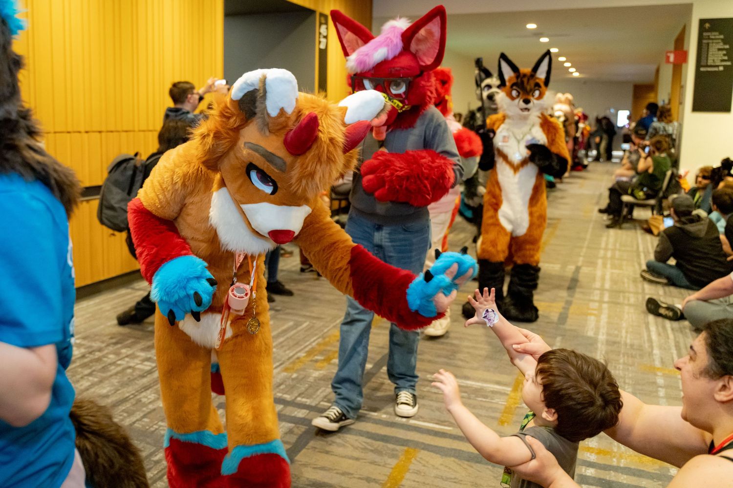 Mascot characters in colorful costumes interact with people at an indoor event in a hallway setting.