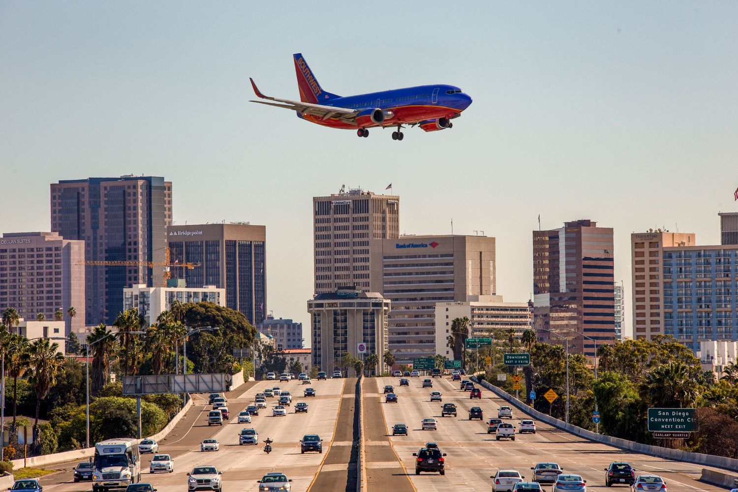 Panoramic cityscape view with commercial airplane, office buildings, and highway traffic during daytime.