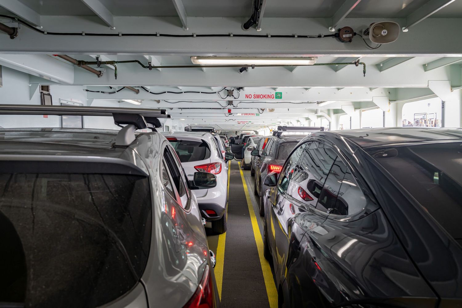 Car deck interior of a ferry showing parked vehicles and overhead mechanical systems.