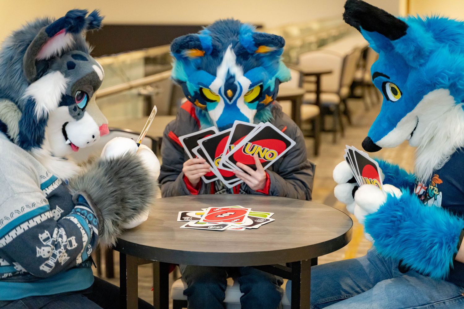 Several fursuit characters playing card games at a convention table with blue and white fur visible in the scene.