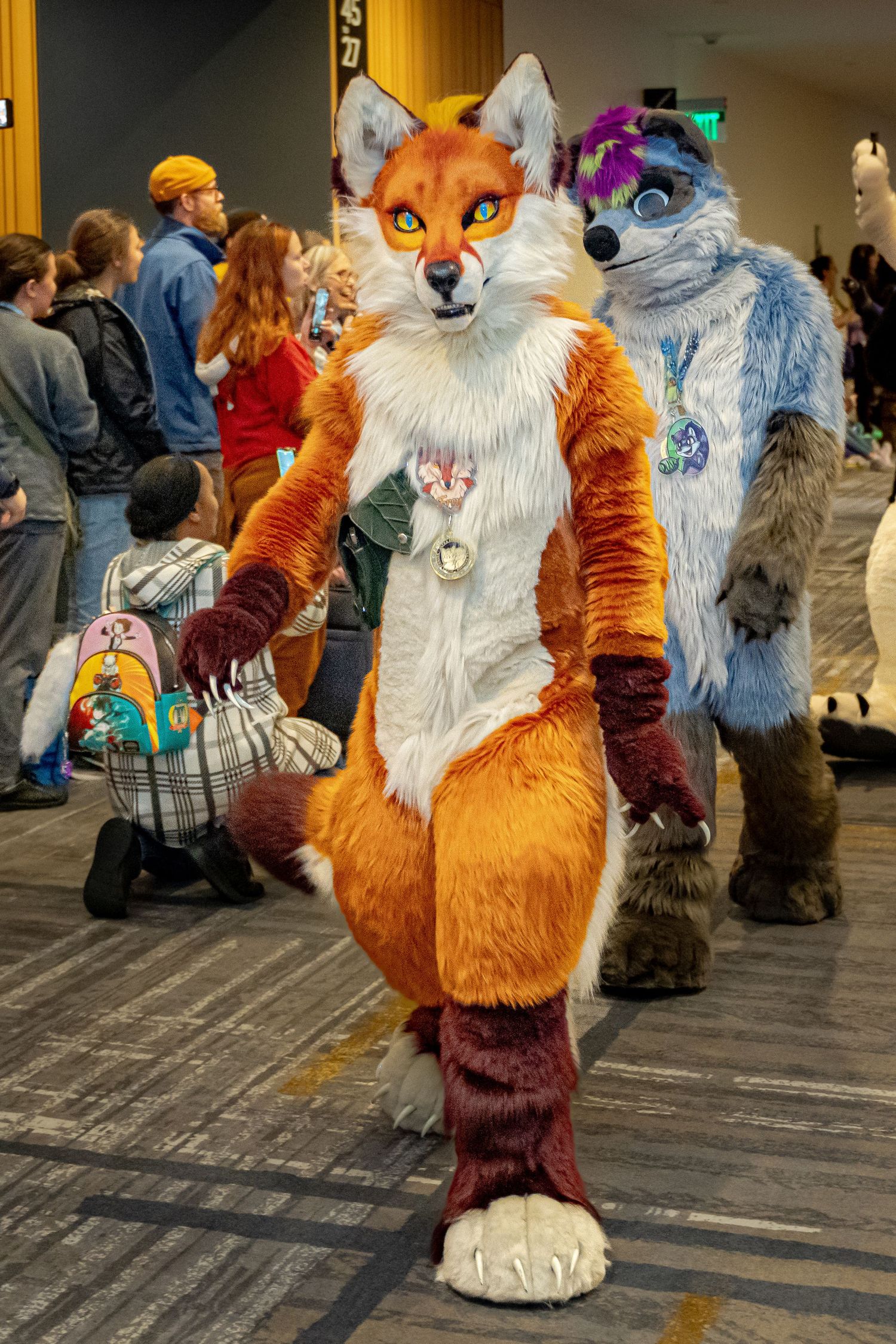 A furry convention attendee in an orange and white fox fursuit poses for photos in a hotel hallway.
