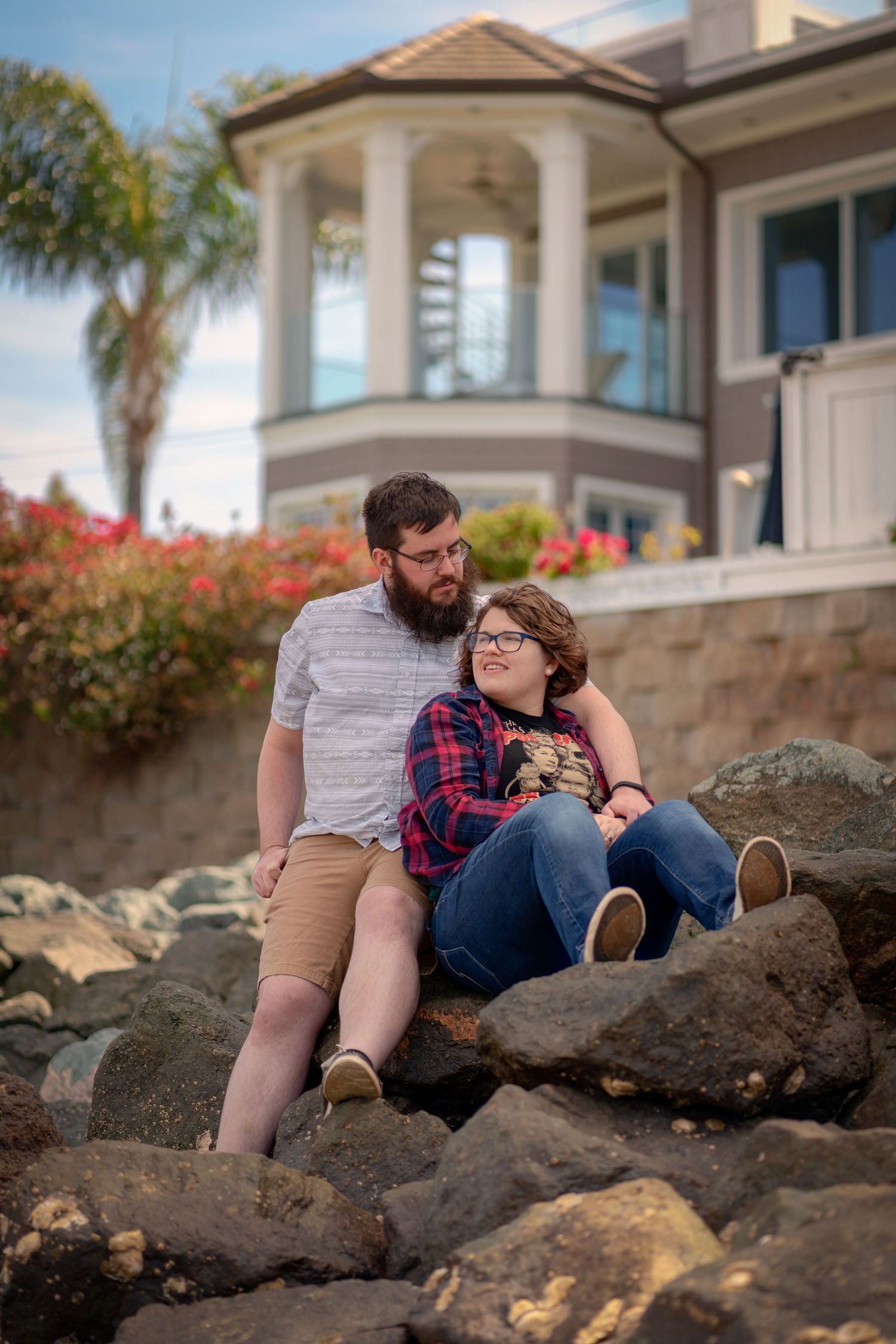 Couple sitting together on rocky coastline with elegant house and palm trees in background.