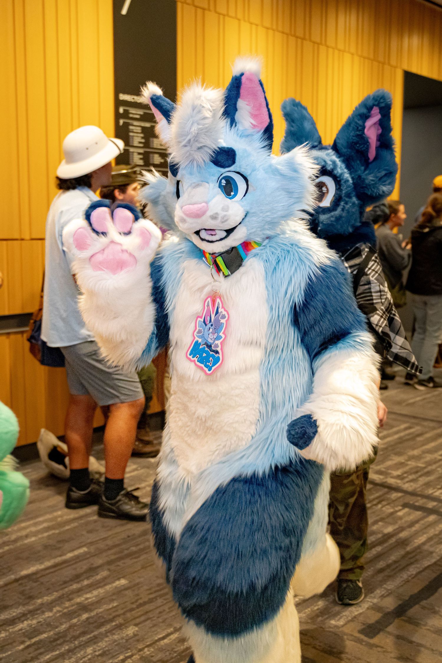 A furry mascot in a blue and white costume with large eyes and paws poses playfully at a convention.