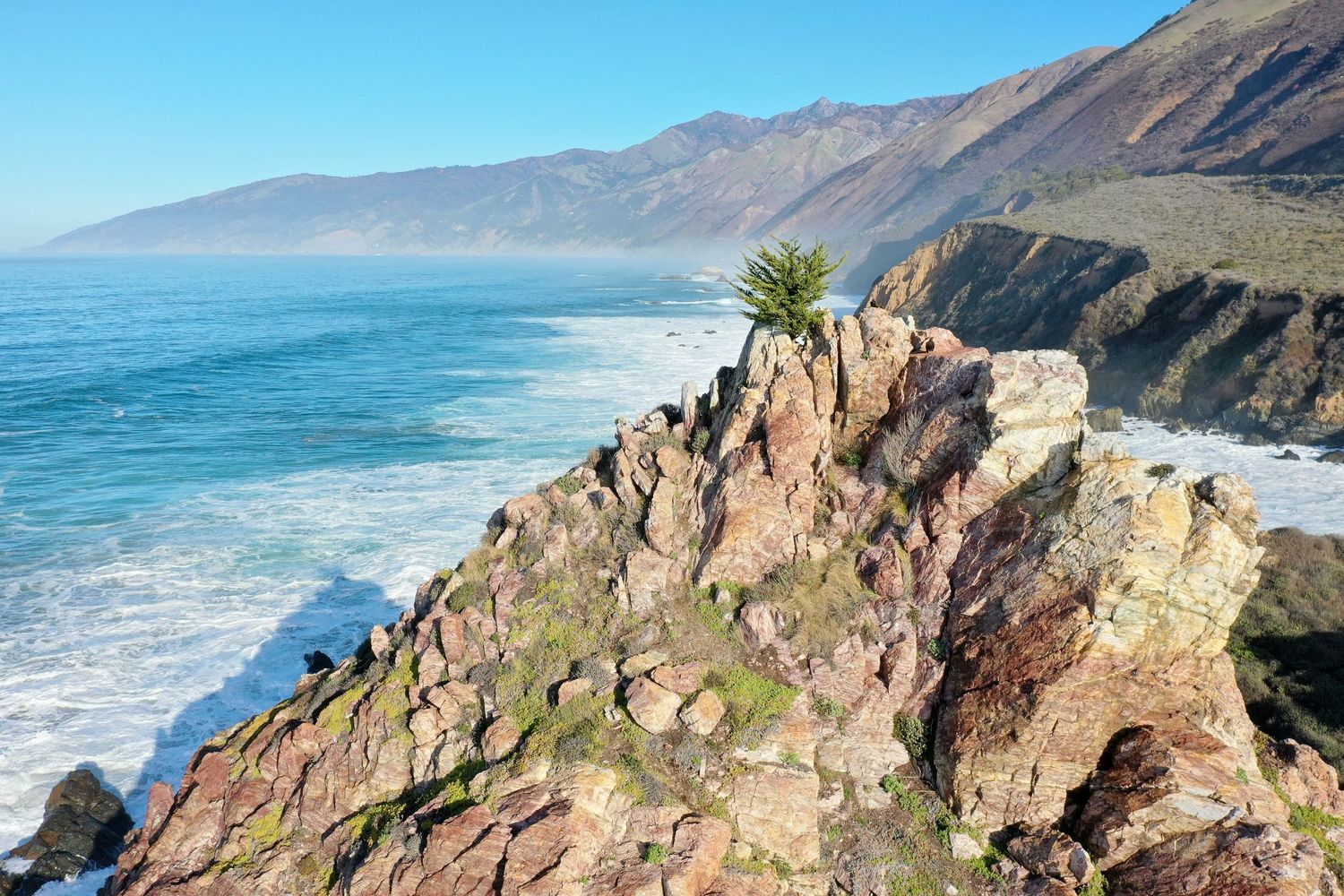 Scenic coastal view of rugged cliffs overlooking the Pacific Ocean with mountains in the background.
