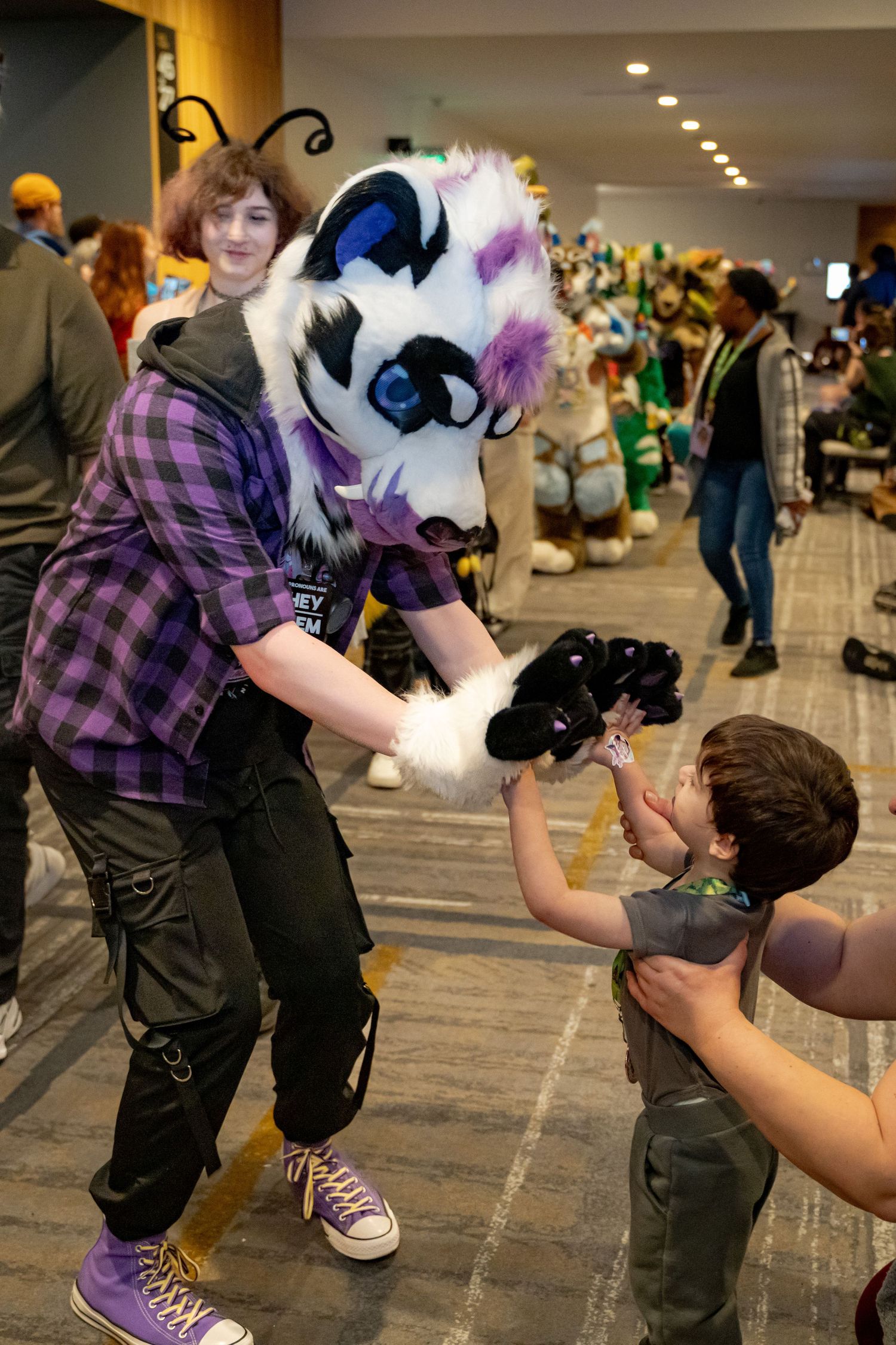 A fursuit character interacts with a person in a convention hall setting while wearing a purple plaid shirt.