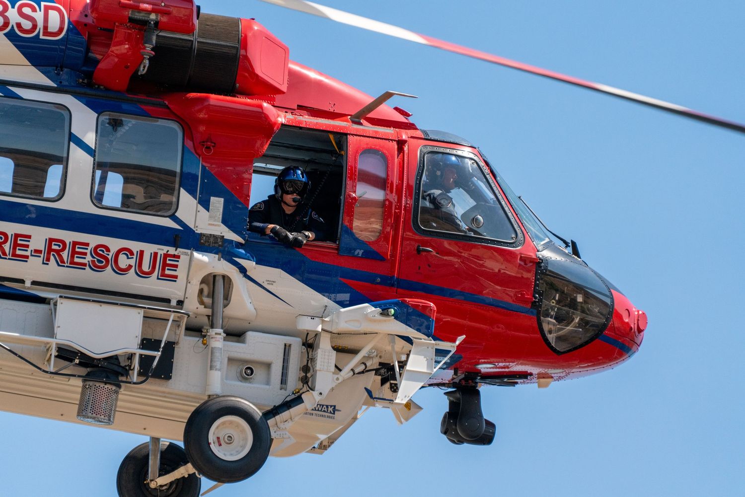 Red rescue helicopter flying against bright blue sky during daytime operations.