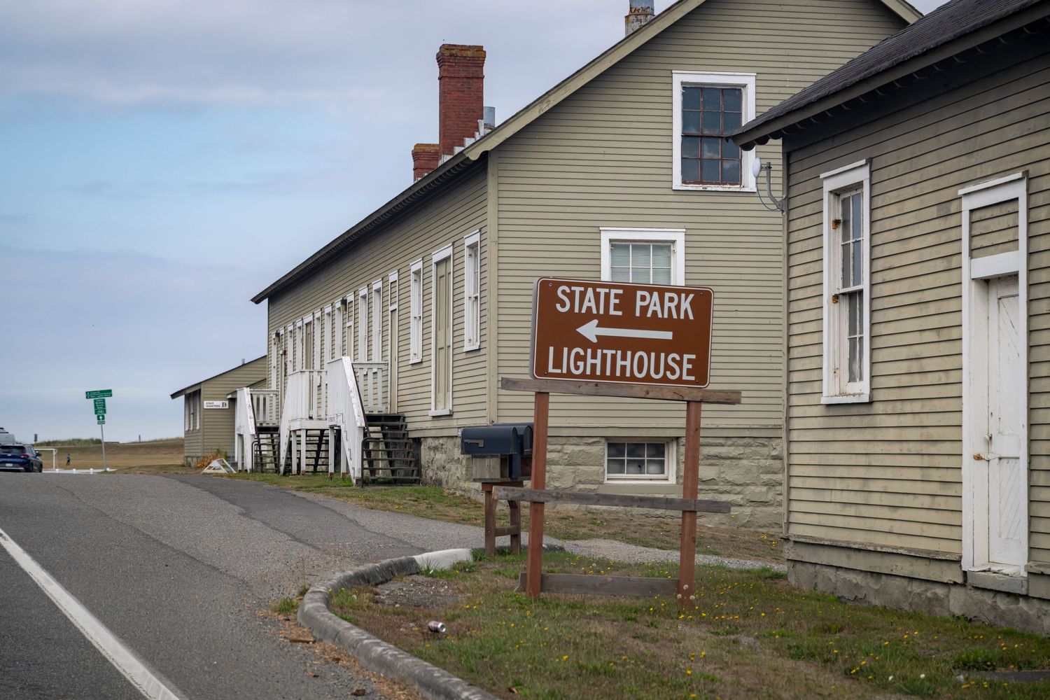 Beige lighthouse building with red brick chimney sits on grassy coastal grounds.