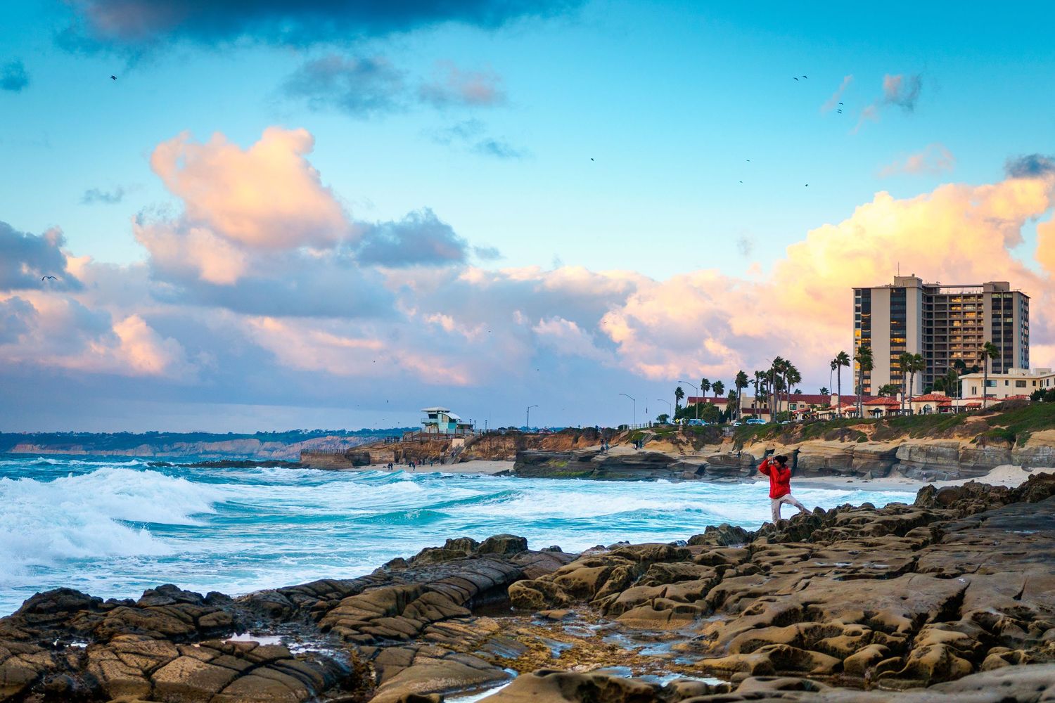 Coastal panorama at sunset showing rocky shoreline and beach with buildings in the distance.