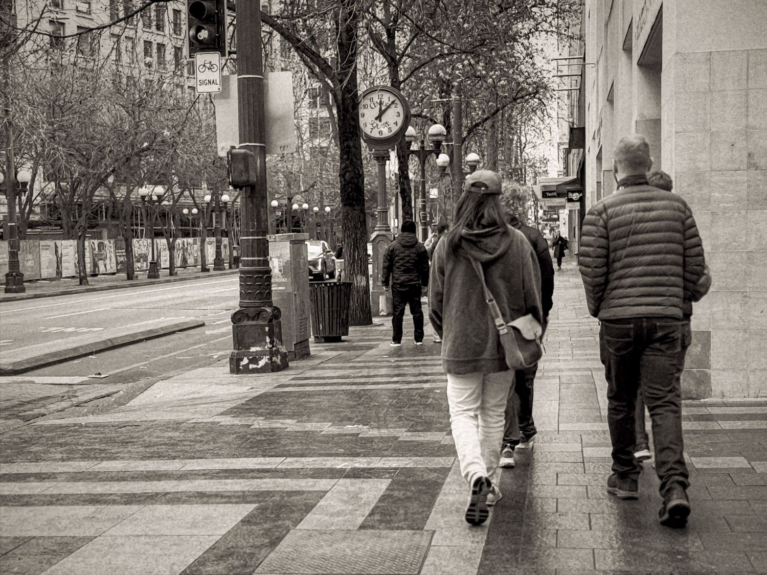 Black and white photograph showing people walking along a tree-lined urban street with historic buildings.