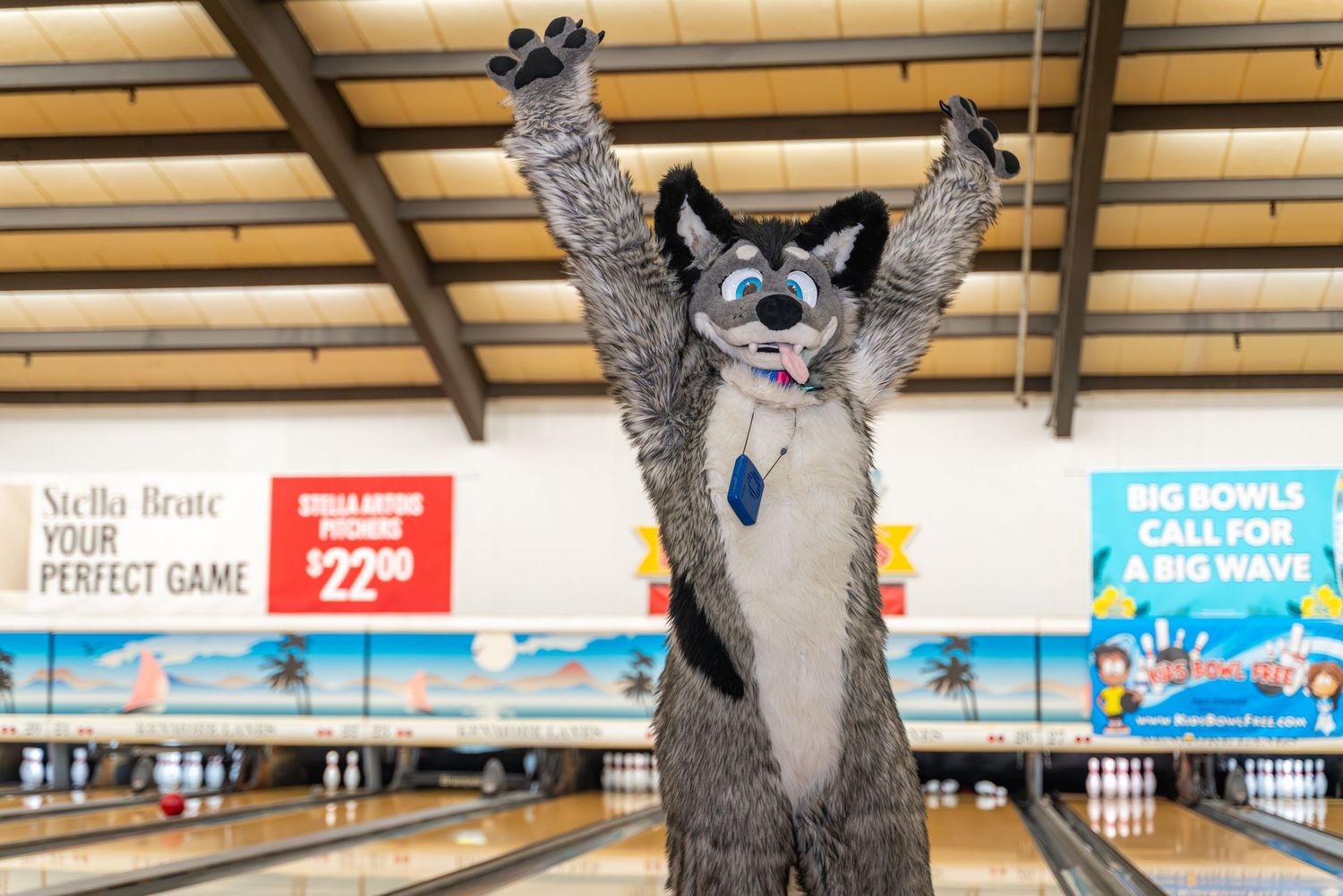 A grey and white fursuit character raises their arms in victory at the bowling alley.