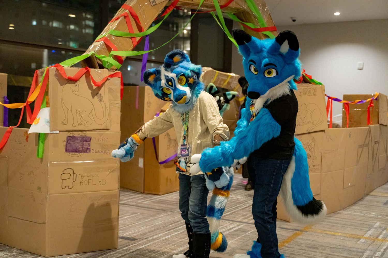 Time-lapse sequence shows a furry blue mascot character moving cardboard boxes in an indoor space with colorful decorations.