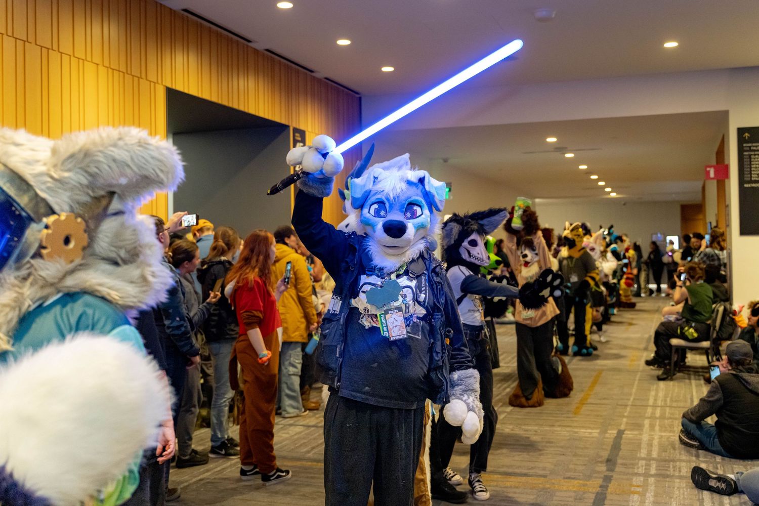 Cosplay convention attendees gather in a hallway with wood paneling and blue lighting for photos and socializing.