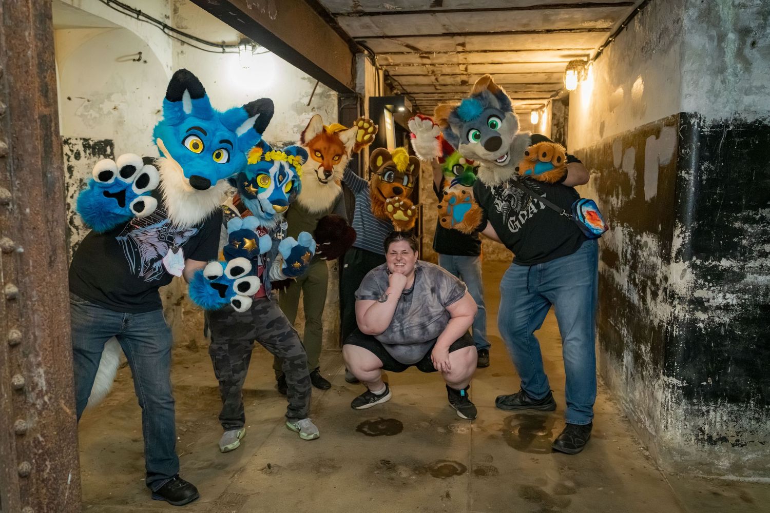 People in colorful fursuit costumes gather in a dimly lit concrete tunnel or hallway.