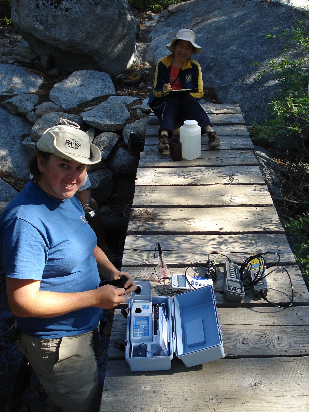 Someone in a blue shirt and sun hat uses scientific equipment on a wooden surface outdoors.