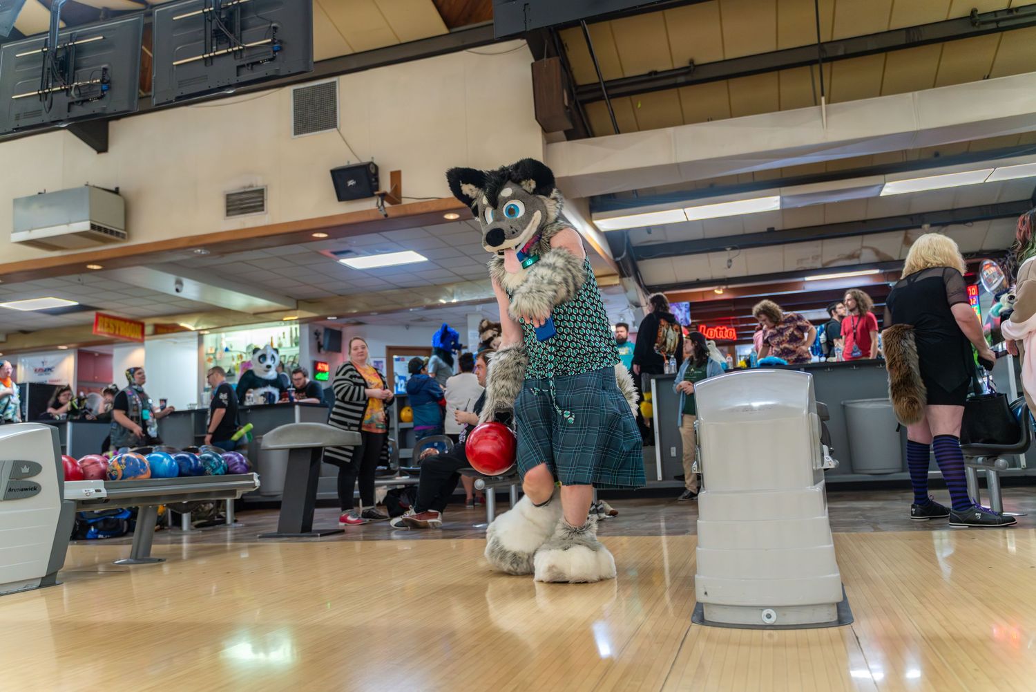 Wide panoramic view of a bowling alley with fursuiters and guests gathered for an event.
