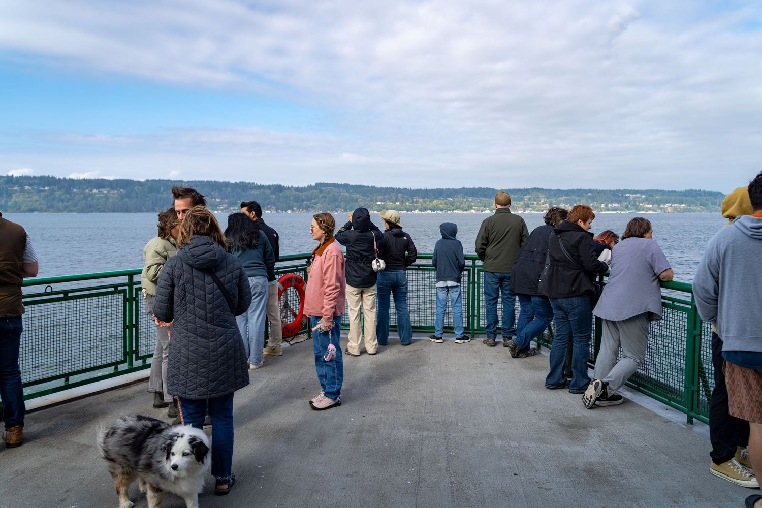 Passengers gather at ferry deck railing overlooking water and distant shoreline.