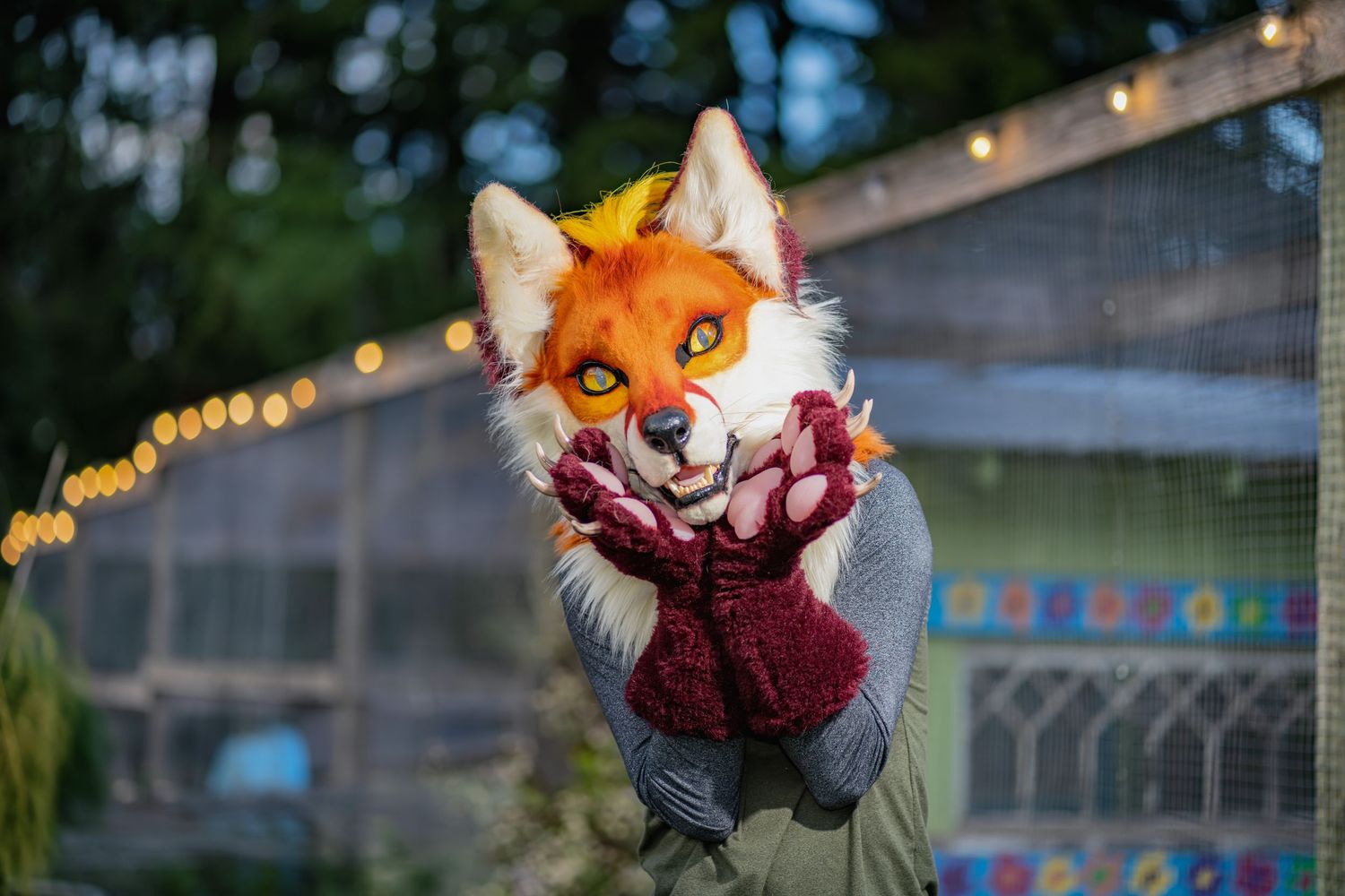 Orange and white fursuit character poses against string lights and garden backdrop.