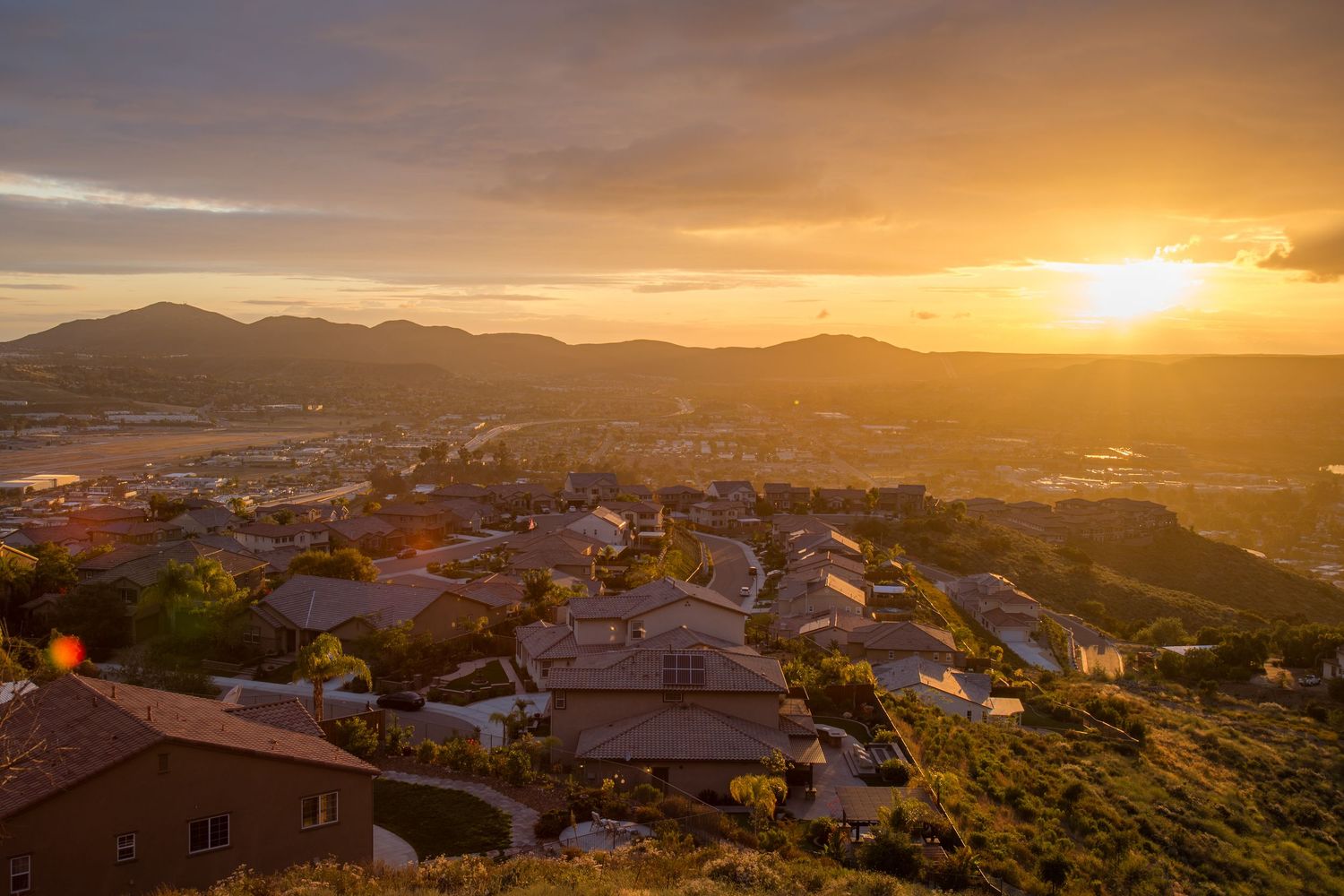 Golden sunset panorama over a hillside residential neighborhood with mountains in the background.