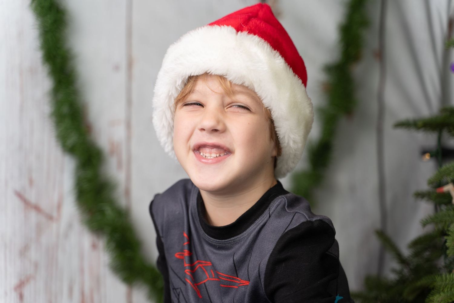 Child wearing red Santa hat and festive clothing in front of holiday decorations.