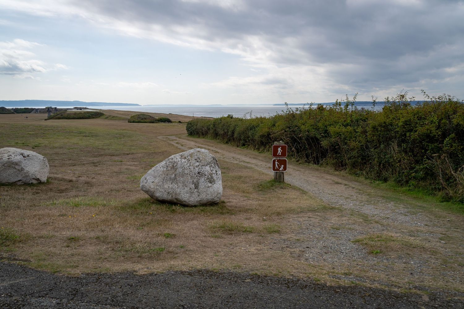 A dirt trail with large boulders leads through coastal terrain under cloudy skies.