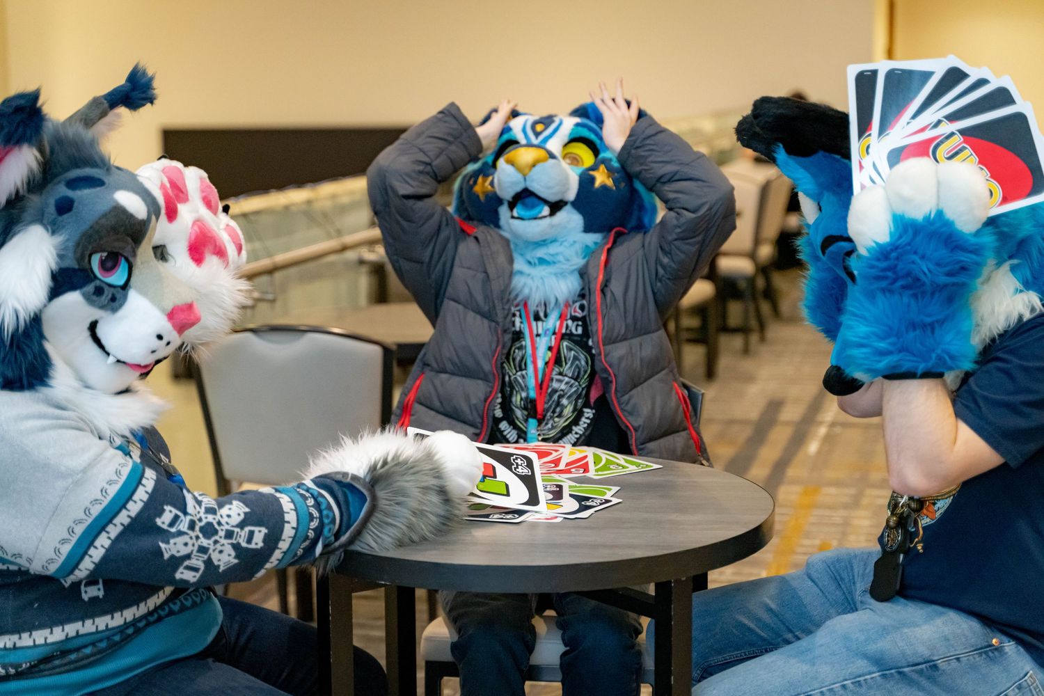 Several furry mascot characters seated at tables in what looks like a convention or meeting space.