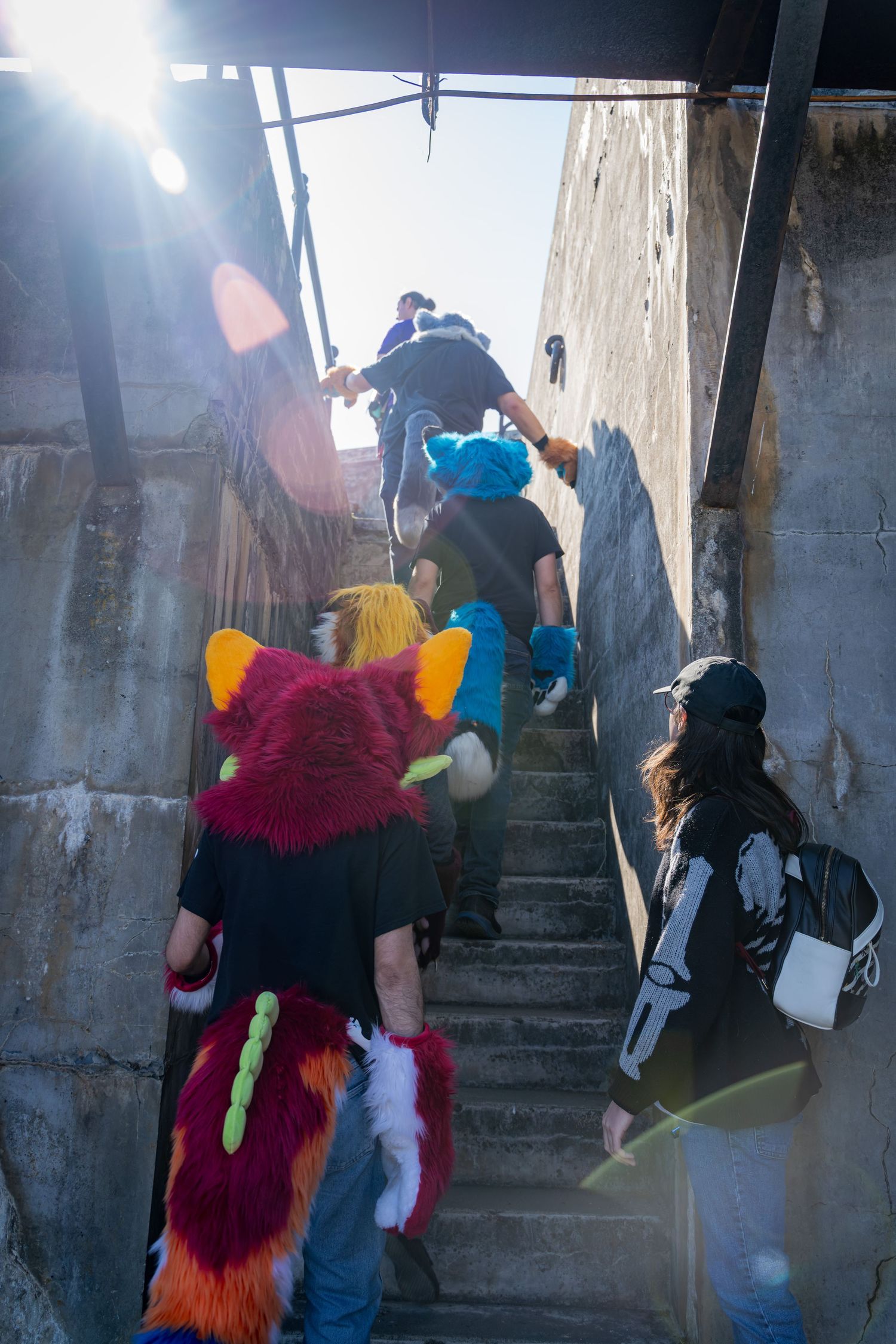 Fursuit performers ascend concrete stairs in a bunker with dramatic lighting streaming in.