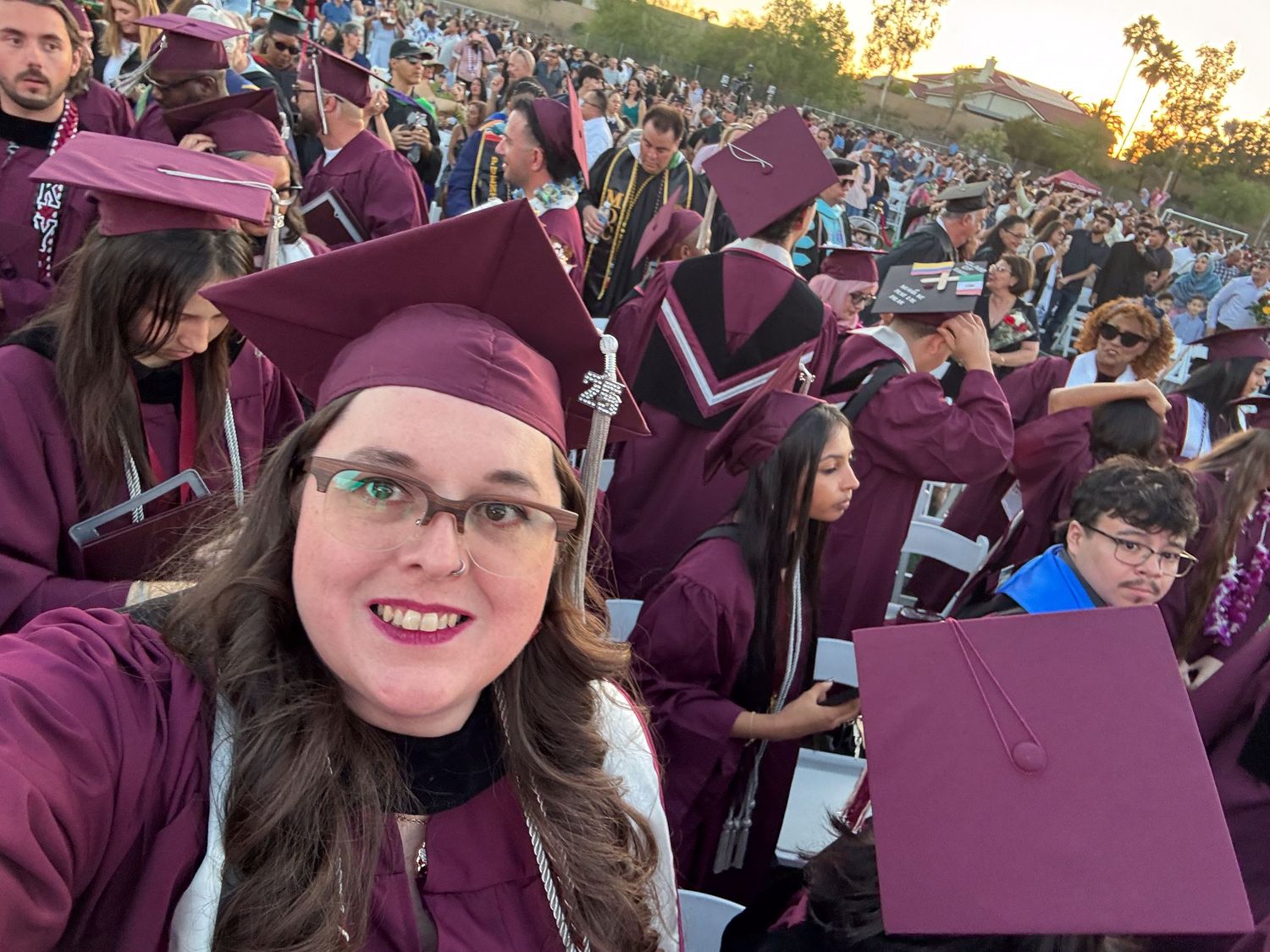 Large crowd wearing maroon graduation caps and gowns gathered outdoors during sunset ceremony.