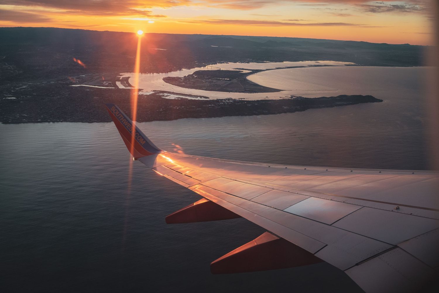 Sunset view from airplane window showing orange sky over coastal landscape.