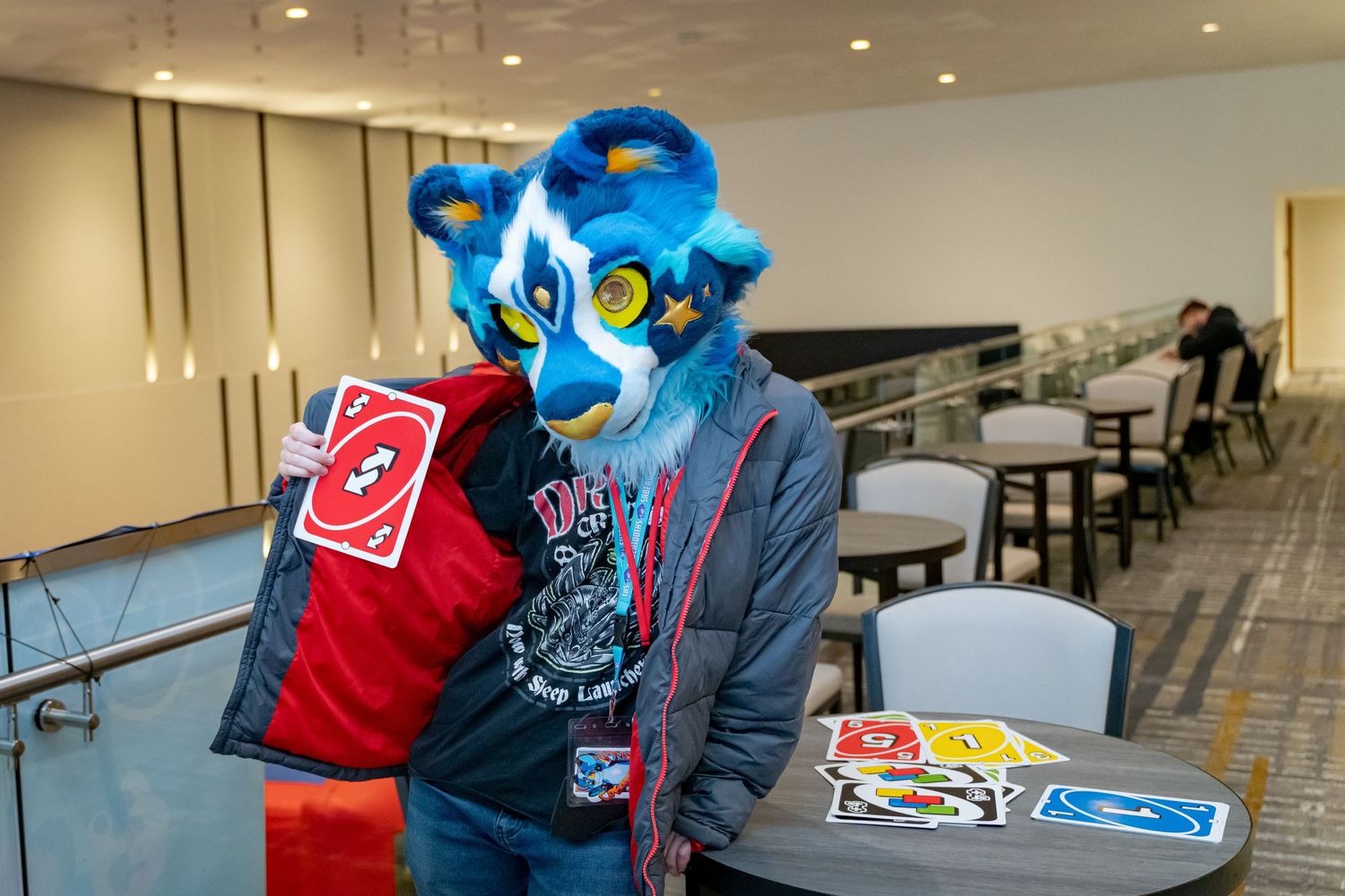 A person in a bright blue fursuit head and casual clothing sits at a table with playing cards.