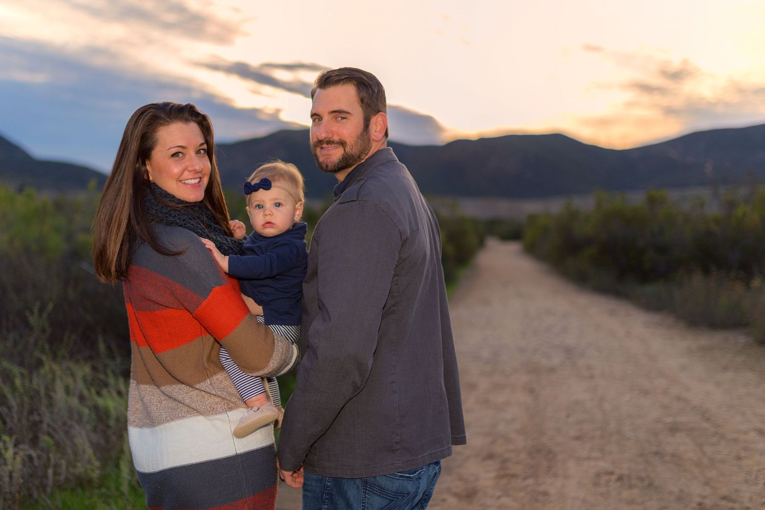 Family standing together on dirt path at sunset with mountains in background.