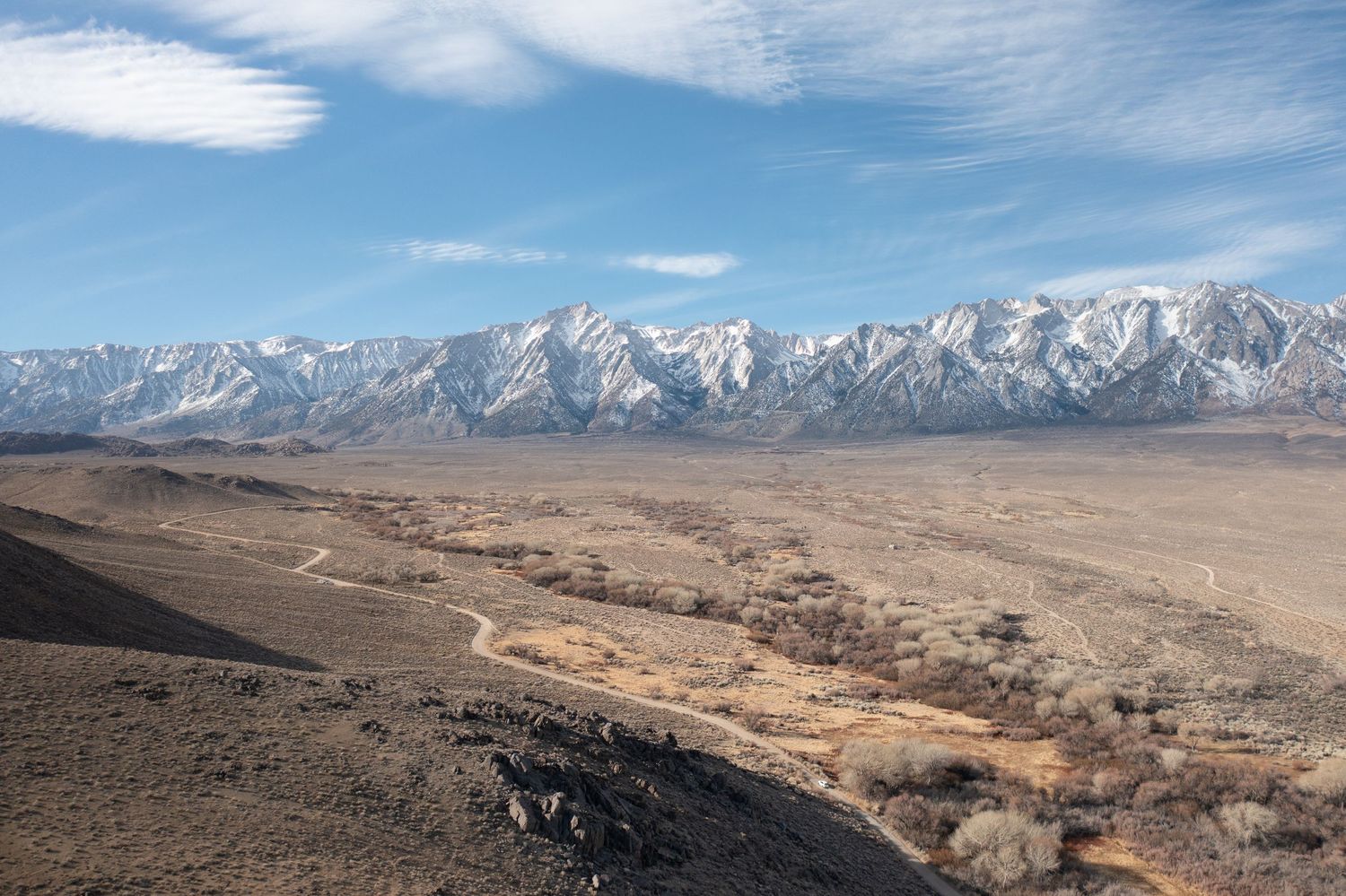 Panoramic mountain landscape with snow-capped peaks and desert valley under cloudy skies.