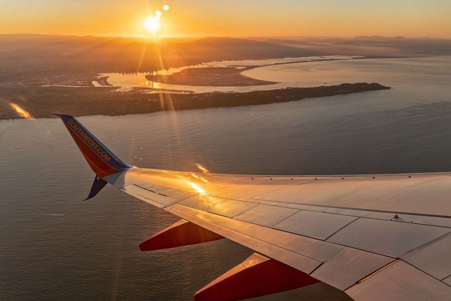 Golden sunset view from airplane window showing wing silhouette over coastal landscape.