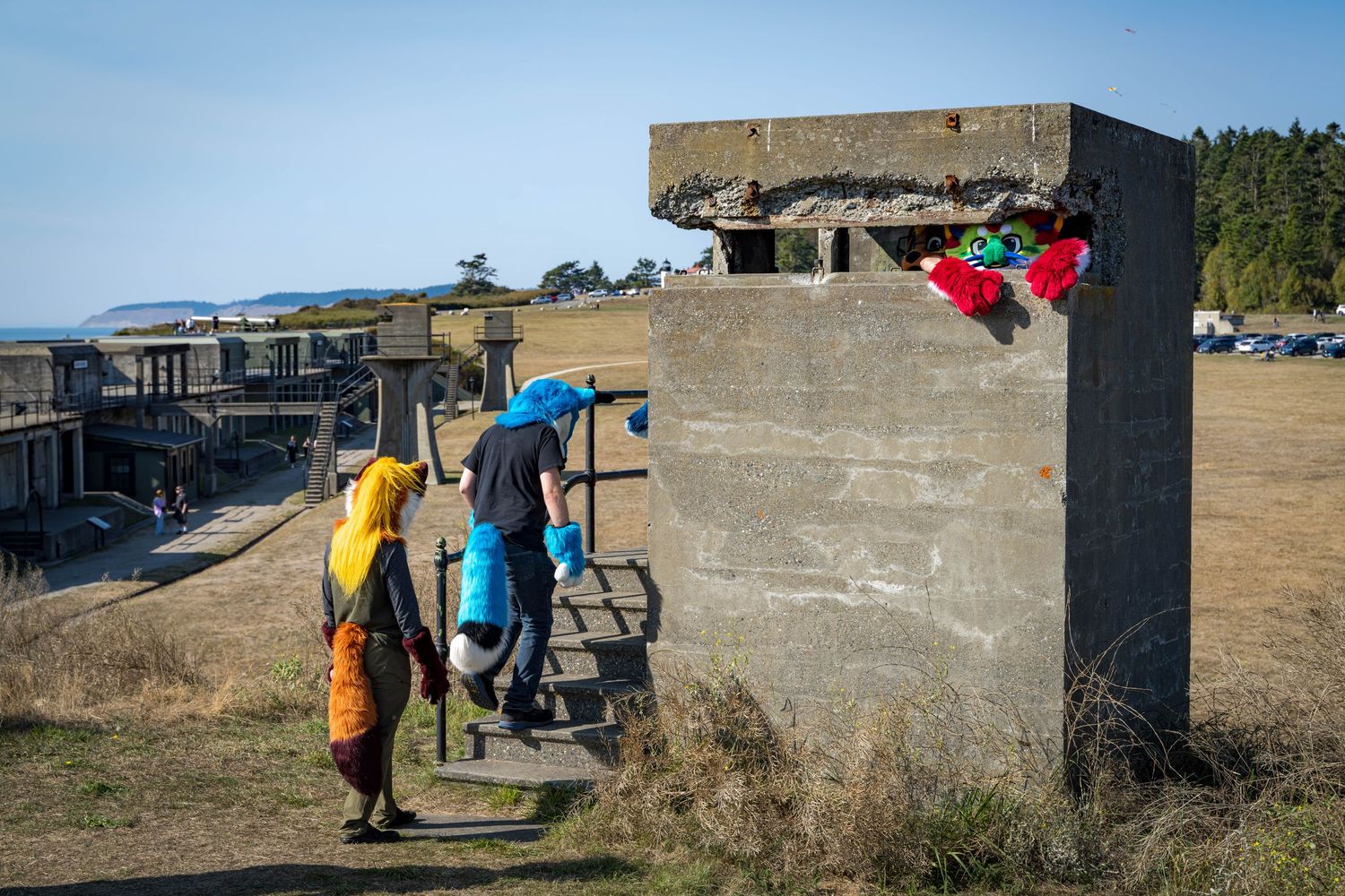 People in colorful outfits walk along a concrete military bunker structure on a sunny day.