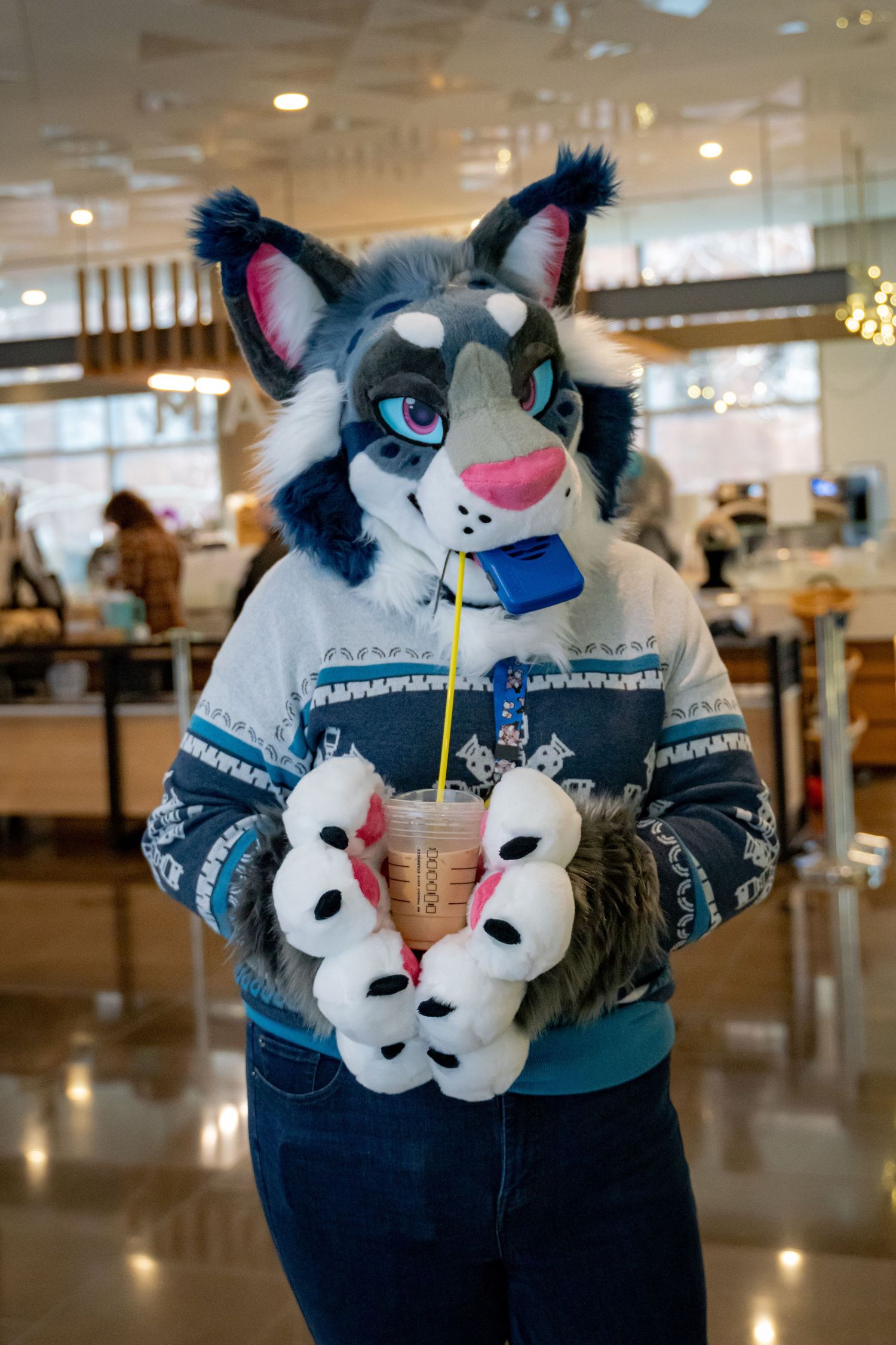 A fursuit character wearing a winter sweater holds an iced coffee drink in a mall food court setting.