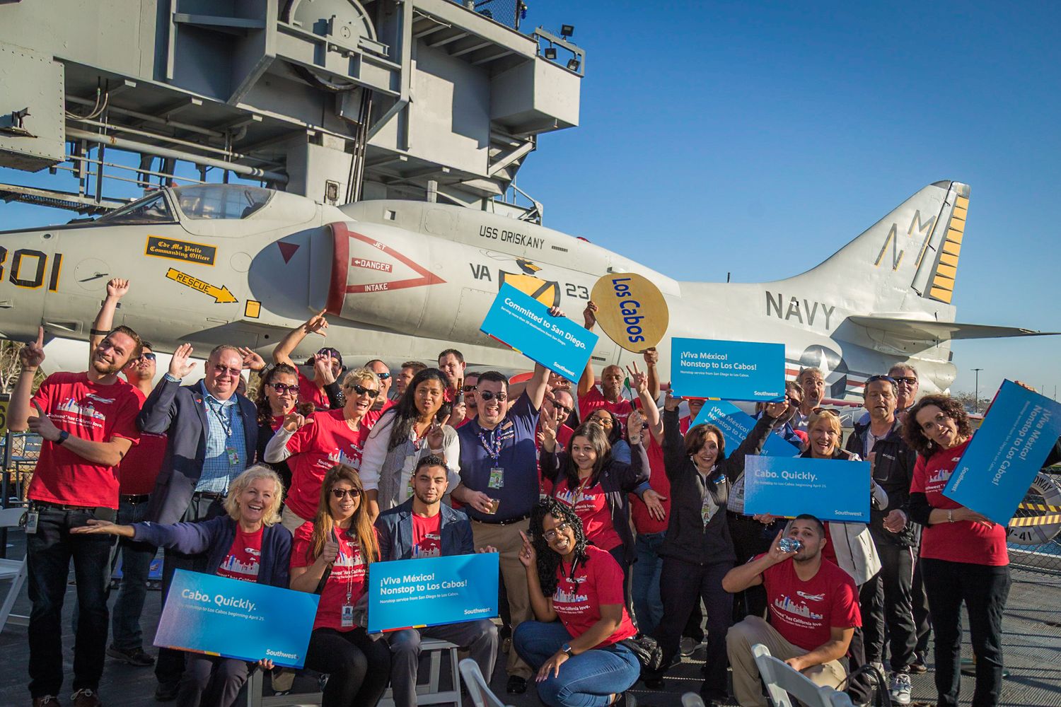 Group of people in red shirts gathered outside near aircraft and military vehicles.