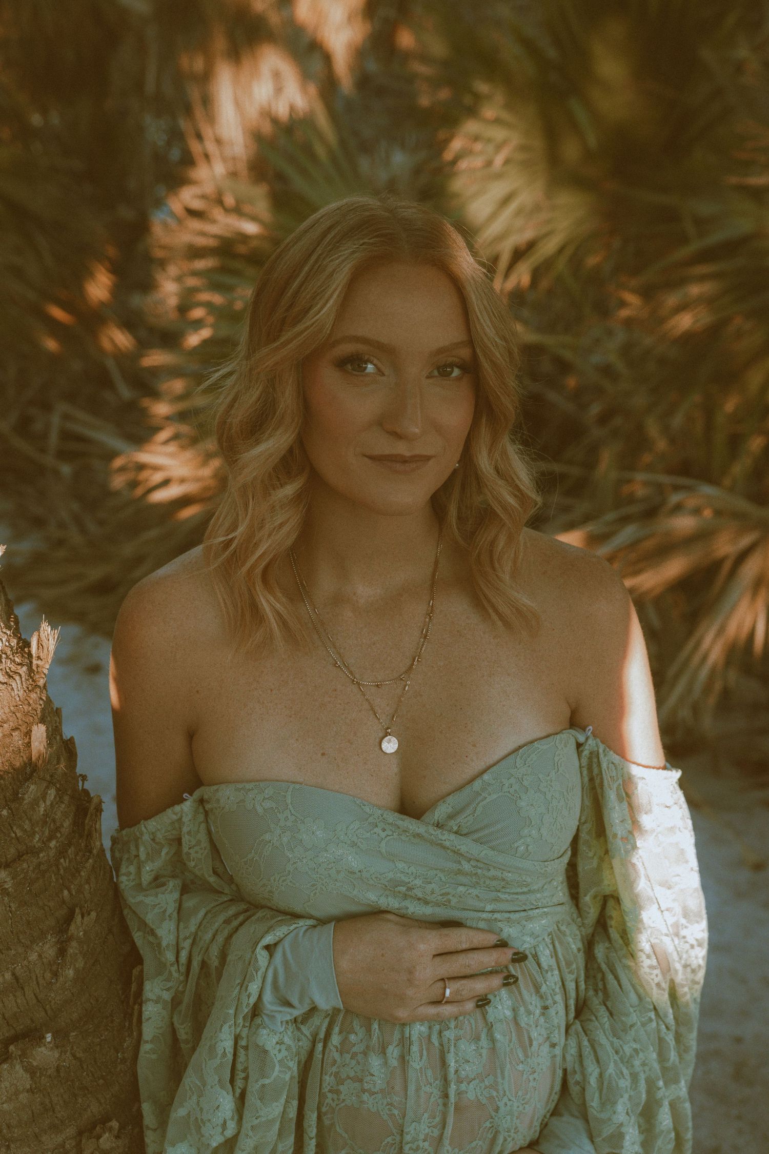 pregnant woman with blonde hair poses with her hand on her belly between palm trees on Caspersen Beach in Venice Florida