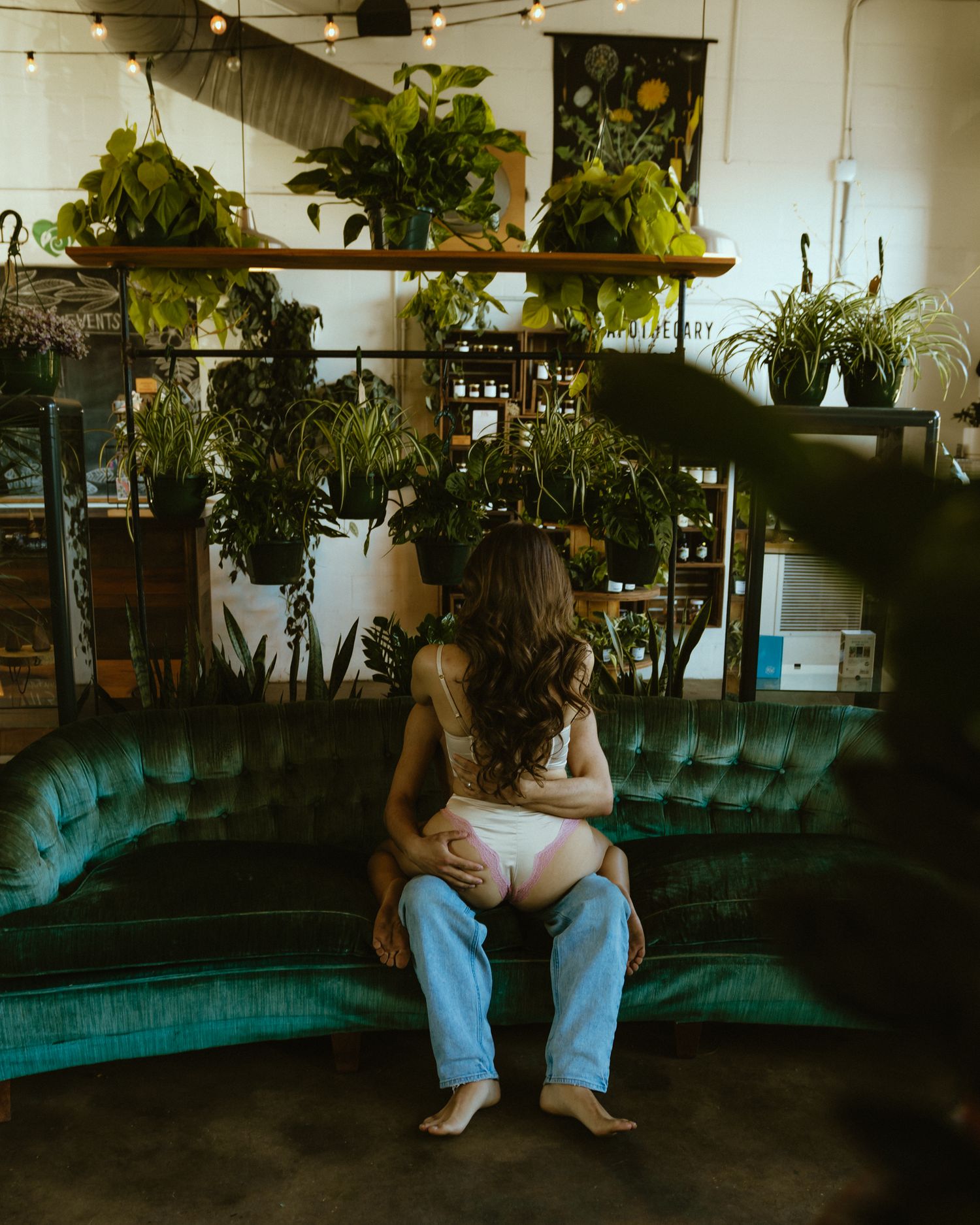 man of the model couple sits facing forward on a green velvet couch. The woman of the model couple is wearing a two piece lingerie set