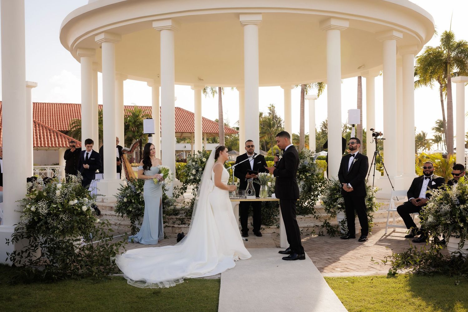 A wedding party poses under elegant white columns and palm trees during an outdoor ceremony at a tropical venue.