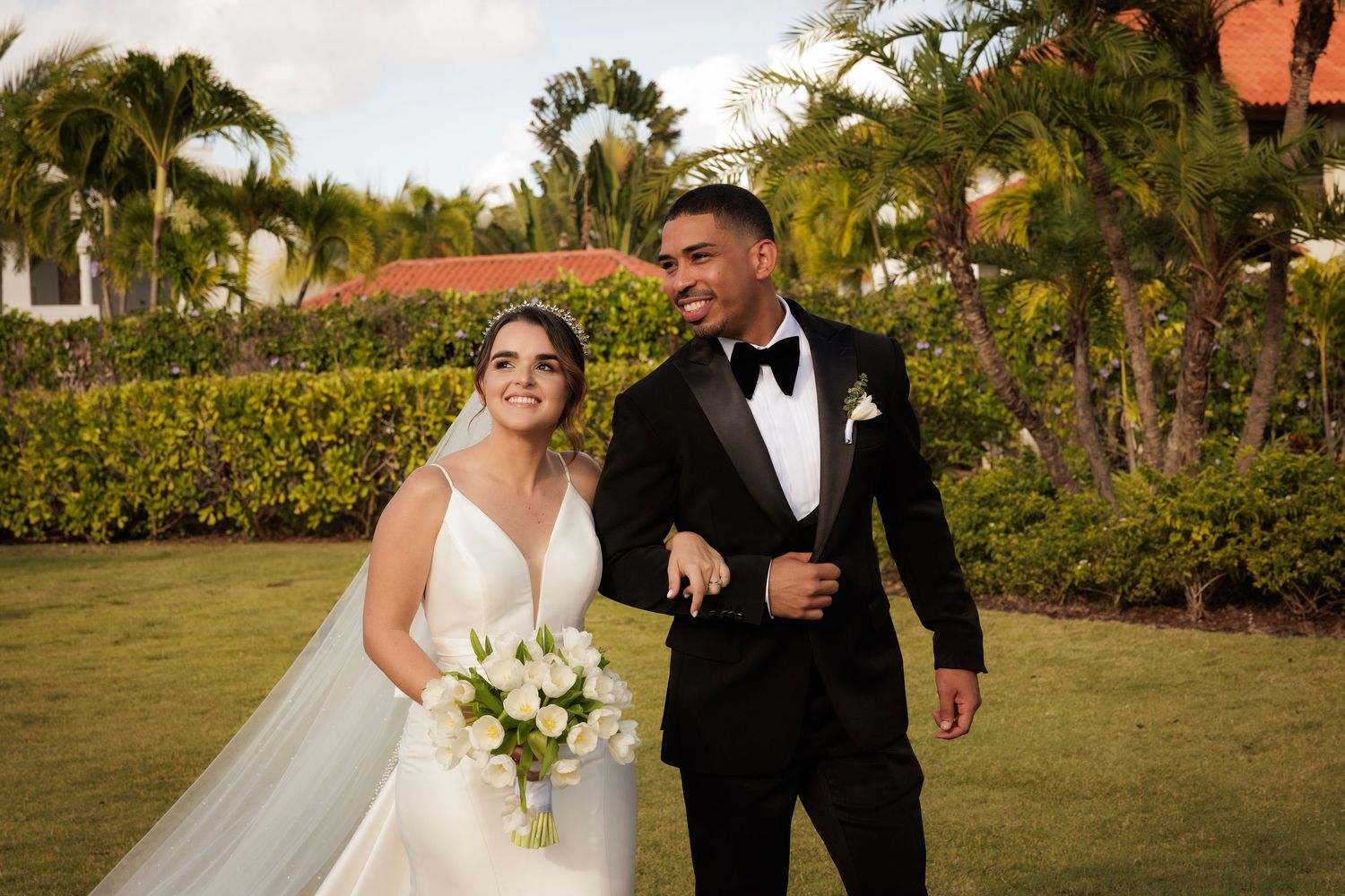 A bride and groom share a romantic moment walking together on lush green grass surrounded by palm trees.