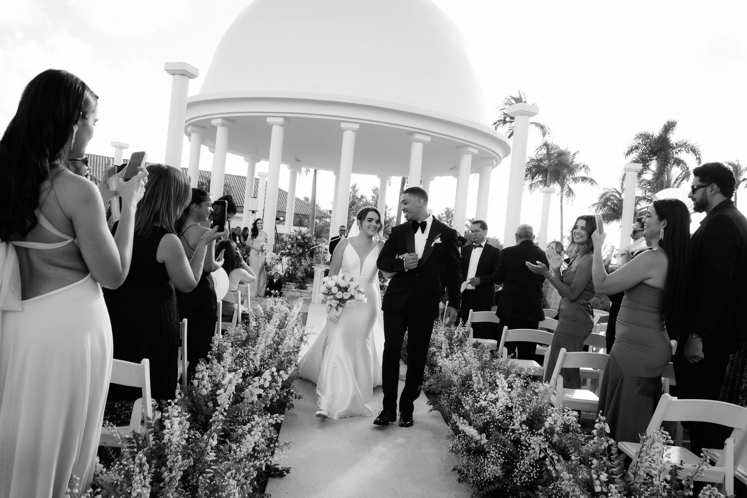 A wedding ceremony takes place under an elegant white pavilion with guests seated and palm trees in the background.