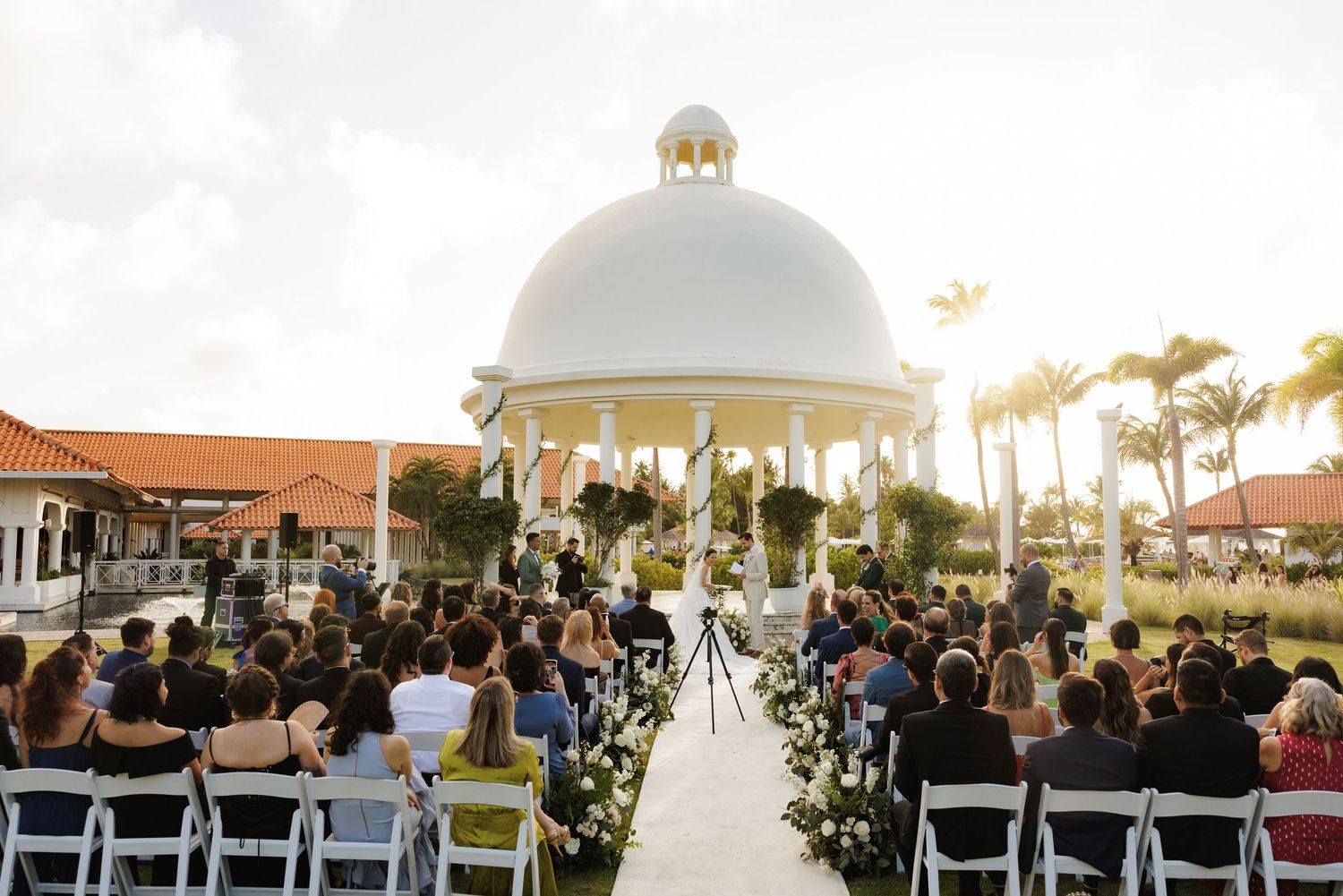 Wedding ceremony guests seated outdoors under palm trees and white gazebos at golden hour near the ocean.