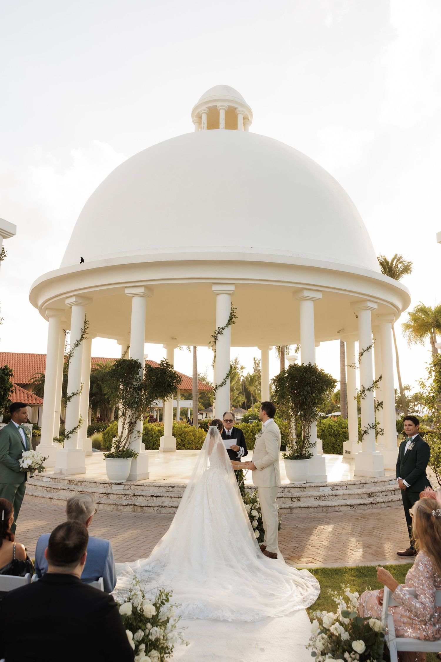 Wedding ceremony under elegant white domed gazebos surrounded by palm trees at sunset.