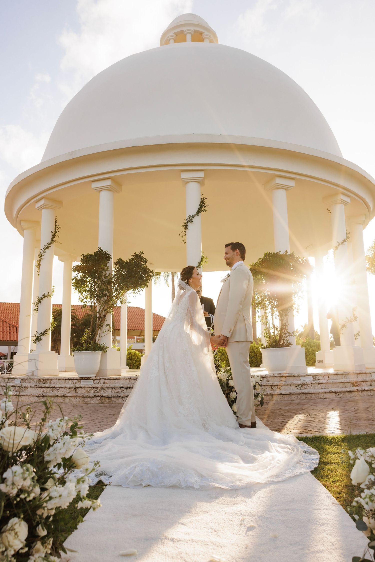Bride in flowing white gown poses under elegant white domed gazebos at sunset during a tropical destination wedding photoshoot.
